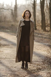 Mature woman with hands in overcoat pockets walking on road at forest - OGF00434