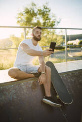 Gut aussehender Mann, der ein Selfie mit seinem Smartphone macht, während er auf einer Sportrampe im Skateboardpark sitzt - RAEF02383