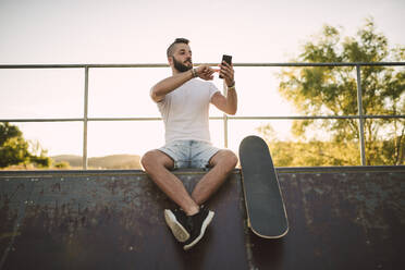 Gut aussehender junger Mann, der sein Smartphone benutzt, während er auf einer Sportrampe im Skateboardpark sitzt - RAEF02384