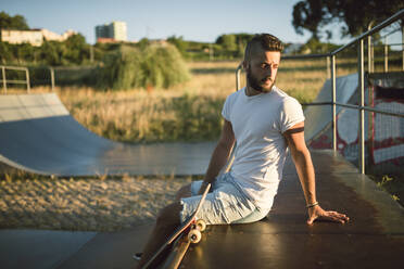 Nachdenklicher junger Mann, der wegschaut, während er auf einer Sportrampe im Skateboardpark sitzt - RAEF02385