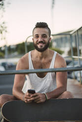 Lächelnder gutaussehender Mann mit Skateboard, der ein Smartphone benutzt, während er gegen den klaren Himmel sitzt - RAEF02387