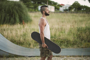 Gut aussehender junger Mann, der ein Skateboard hält und wegschaut, während er an Land steht - RAEF02388