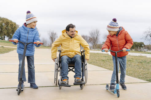 Fröhliche Jungen fahren Roller, während der Vater im Rollstuhl auf dem Gehweg im Park sitzt - JCZF00107