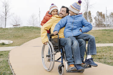 Happy man with playful sons riding wheelchair on footpath at park - JCZF00108