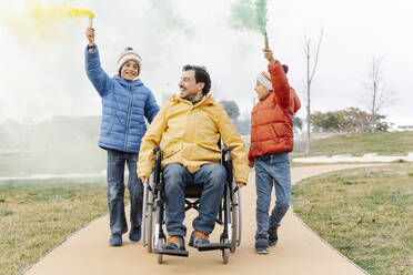 Mann fährt Rollstuhl, während Jungen mit Leuchtrakete auf Fußweg in Park gehen - JCZF00111