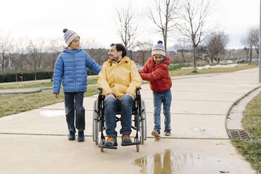 Söhne unterhalten sich und helfen ihrem im Rollstuhl sitzenden Vater im Park - JCZF00118