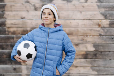 Thoughtful boy wearing warm clothing holding soccer ball while standing against wall - JCZF00119