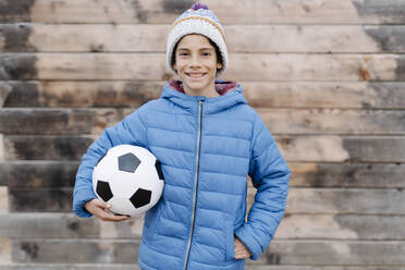 Lächelnder Junge in warmer Kleidung, der einen Fußball hält und an der Wand steht - JCZF00120