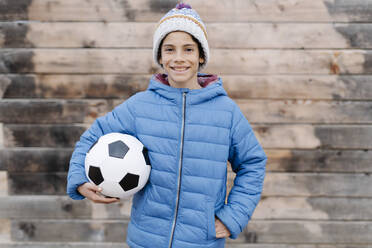 Smiling boy wearing warm clothing holding soccer ball while standing against wall - JCZF00120