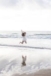 Carefree young woman with arms raised jumping at shore against sky - FVSF00376