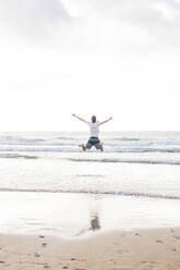 Carefree young man with arms outstretched jumping at beach against sky - FVSF00379