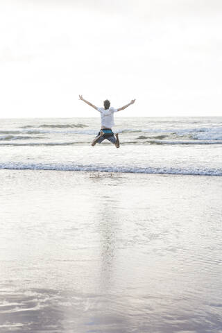 Unbekümmerter junger Mann mit ausgestreckten Armen, der gegen den klaren Himmel ans Ufer springt, lizenzfreies Stockfoto