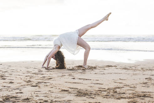 Junge Frau in weißem Kleid tanzt auf Sand am Strand gegen klaren Himmel - FVSF00382