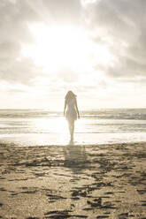 Junge Frau spaziert am Strand gegen den bewölkten Himmel bei Sonnenuntergang - FVSF00388