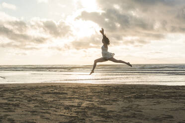 Happy young woman with arms raised jumping at beach against cloudy sky during sunset - FVSF00392