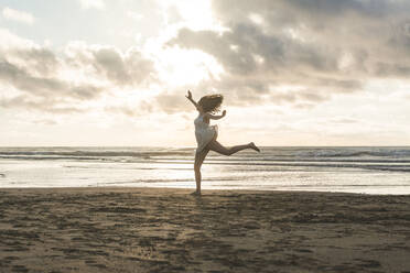 Unbeschwerte junge Frau springt am Strand gegen bewölkten Himmel bei Sonnenuntergang - FVSF00393