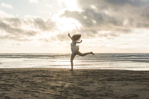 Unbeschwerte junge Frau springt am Strand gegen bewölkten Himmel bei Sonnenuntergang - FVSF00393