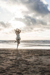 Carefree woman with arms raised dancing at beach against cloudy sky during sunset - FVSF00394