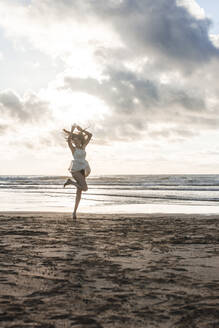 Sorglose Frau mit erhobenen Armen tanzt am Strand gegen den bewölkten Himmel bei Sonnenuntergang - FVSF00394