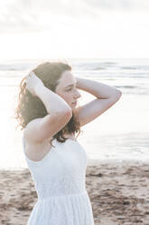 Nachdenkliche junge Frau in weißem Kleid mit Händen im Haar am Strand stehend - FVSF00396
