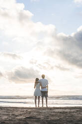 Loving young couple with arms around looking at sea against cloudy sky - FVSF00397