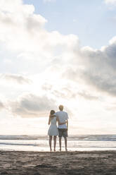 Verliebtes junges Paar, das sich in den Armen liegt und auf das Meer vor einem bewölkten Himmel blickt - FVSF00397