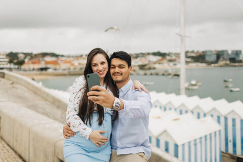 Expectant couple taking selfie through smart phone at harbor, Cascais, Portugal stock photo