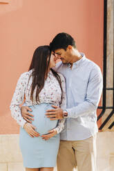Romantisches schwangeres Paar, das mit den Händen auf dem Bauch vor einer rosa Wand in der Stadt steht - DCRF00258