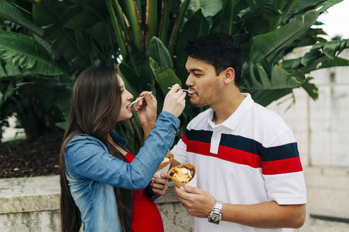 Expectant couple feeding ice creams to each other during sunny day - DCRF00262