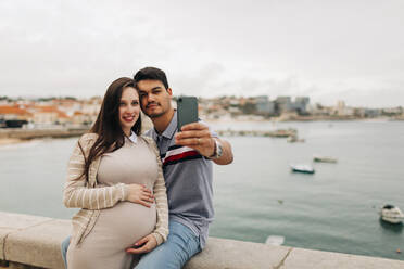 Ein schwangeres Paar macht ein Selfie mit seinem Smartphone am Hafen, Cascais, Portugal - DCRF00273