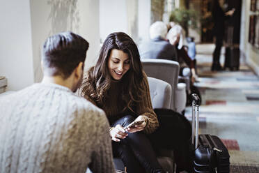 Lächelnde Frau, die ein Smartphone benutzt, während sie mit einem männlichen Partner auf einem Stuhl im Hotel sitzt - MASF18386