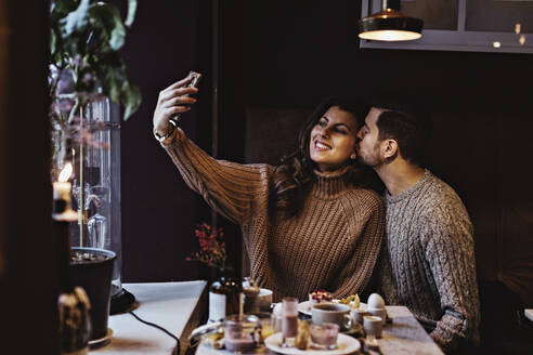 Lächelnde Frau, die ein Selfie über ihr Smartphone macht, während ihr männlicher Partner sie im Hotel küsst - MASF18396