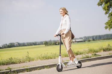 Glückliche Geschäftsfrau, die bei sonnigem Wetter mit einem Elektroroller unterwegs ist - UUF20497