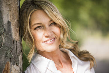 Thoughtful mature businesswoman smiling while leaning on tree trunk during sunny day - UUF20504