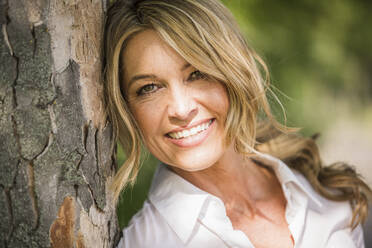 Happy mature businesswoman leaning on tree trunk during sunny day - UUF20505