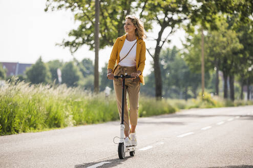 Ältere Frau fährt elektrischen Roller auf der Straße an einem sonnigen Tag - UUF20530