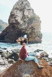 Unbekümmerte junge Frau schüttelt den Kopf, während sie auf einem Felsen am Strand von Ursa sitzt, Portugal - FVSF00446