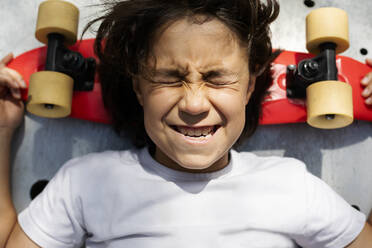 Nahaufnahme eines Jungen mit geschlossenen Augen, der ein Gesicht macht, während er auf einem Skateboard liegt - VABF02976