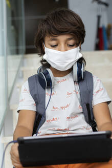 Close-up of boy wearing face mask using digital tablet while sitting on steps in school - VABF02983