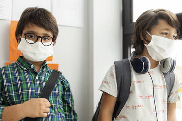 Boy wearing mask standing with friend against wall in school - VABF02987