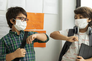 Boys wearing masks giving elbow bump while standing against wall in school - VABF02988