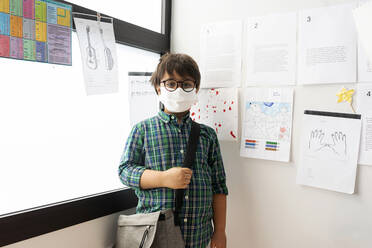 Boy wearing mask with bag standing against wall and window in school - VABF02996