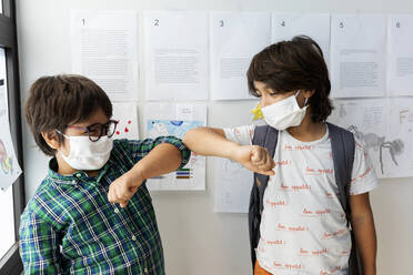 Schoolboys wearing masks giving elbow bump while standing against wall in school - VABF03000