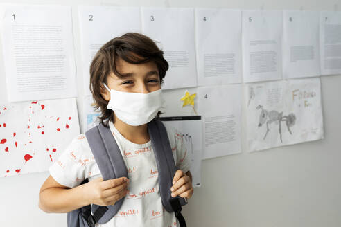 Boy wearing mask standing against papers stuck on wall in school - VABF03001