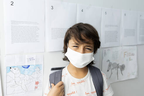 Close-up of boy wearing mask standing against papers stuck on wall in school - VABF03005