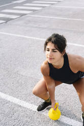 Confident young woman lifting kettlebell while standing on road - JCMF00789