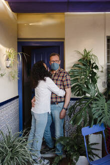 Romantic couple wearing masks embracing while standing against house in yard - AGGF00074