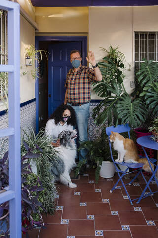 Mann mit Maske winkt mit der Hand, während Frau Hund im Hof streichelt, lizenzfreies Stockfoto