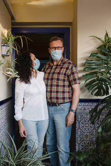 Couple wearing masks standing against house entrance - AGGF00079