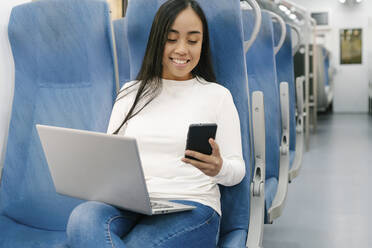 Smiling woman with laptop using smart phone while sitting on seat in train - JCZF00124